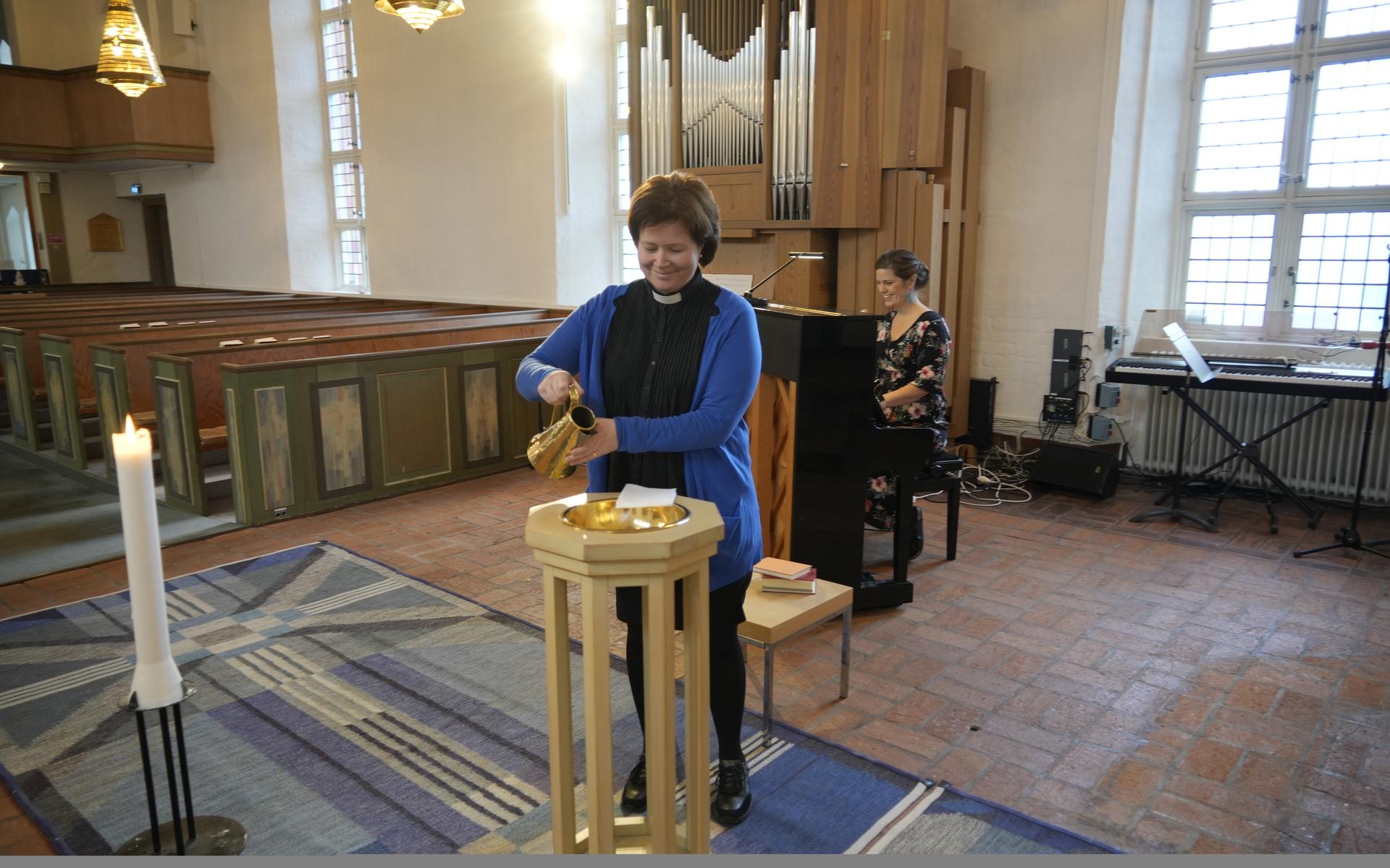 Sofia Eriksson, präst och församlingsherde, häller upp lite vatten i dopfunten. Vid pianot sitter Hannah Magnesköld och värmer upp inför de sånger och psalmer som ska spelas. 